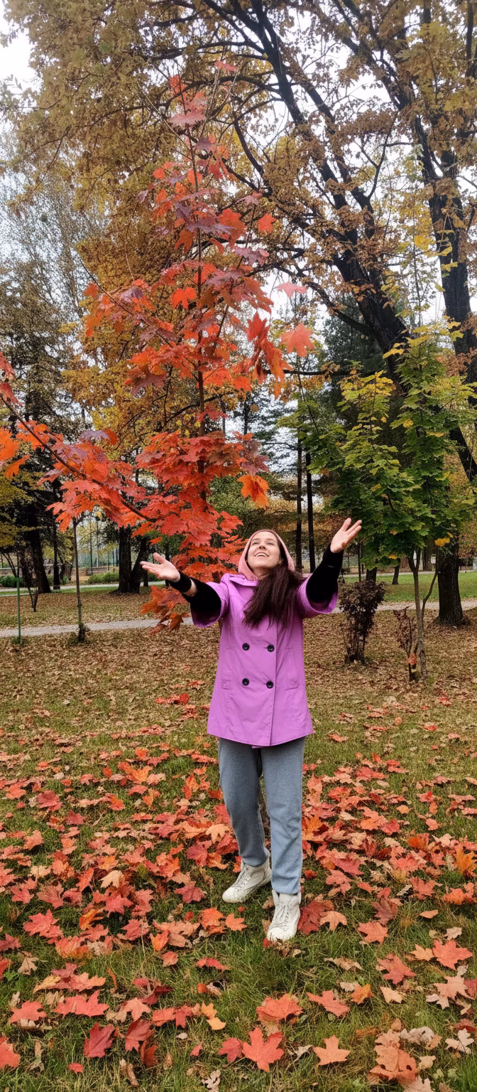 Лариса Примоченко – A joyful young woman with long dark hair, wearing a bright pink-purple hooded coat, light gray jeans, and white sneak…